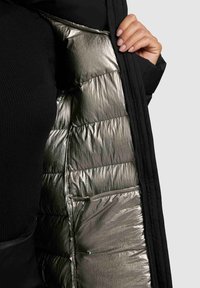 Musta täitemantel, millel on läikiv metallik hõbedane vooder. Näha on vatitekk, millel on sile tekstuur ja timmitud disain.