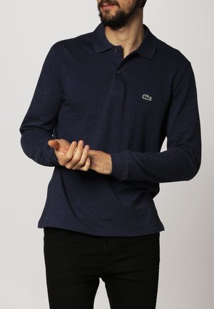 Homme portant un polo bleu marine à manches longues avec logo crocodile, ajustant son poignet droit avec la main gauche sur un fond uni.