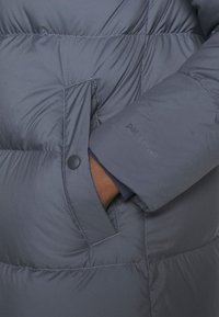 La veste matelassée grise présente un design rembourré, une poche latérale avec un bouton-pression et un texte en relief sur la manche. Tissu lisse et léger.