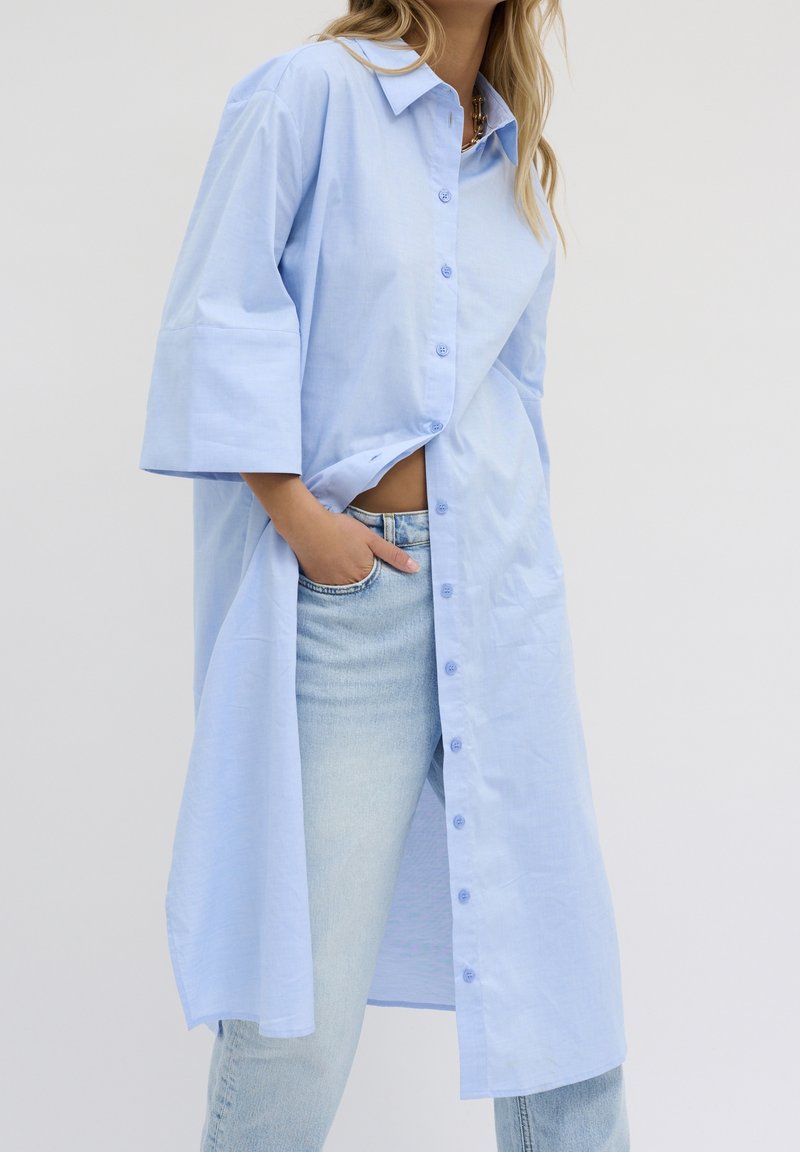 Chemise bleu clair oversize à manches courtes, avec des boutons sur le devant et une coupe droite, associée à un jean en denim clair.