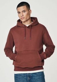 Sudadera burdeos con bolsillo canguro en el frente, capucha con cordón y puños acanalados. Usada sobre una camiseta blanca, complementada con jeans azules.