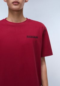 Camiseta de algodón carmesí con cuello redondo clásico, que presenta un logotipo negro impreso en la zona izquierda del pecho. Textura suave, corte relajado.