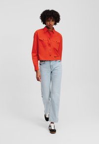 Rode cropped overhemd met knopen en twee borstzakken, gedragen met lichtblauwe hooggerate jeans en zwarte sneakers met witte veters.