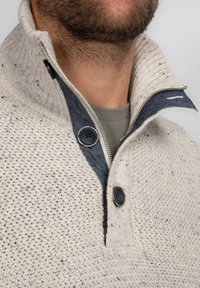 Pull-over en maille beige clair avec une texture mouchetée. Il présente un col bleu marine, une fermeture éclair sur le devant et deux accents de boutons près du cou.