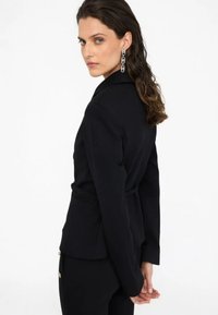 Blazer negro ajustado con mangas largas, cinturón en la cintura y detalles de costura sutiles. Textura suave, bolsillos frontales y diseño de cuello clásico.