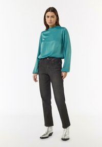 Türkisfarbene Satinbluse mit hohem Kragen und langen Ärmeln, kombiniert mit dunkelblauen Jeans und metallisch-silbernen Stiefeletten.