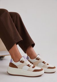 Białe i beżowe, masywne sneakersy z brązowymi zamszowymi akcentami, fioletowym detalem na pięcie i teksturowaną, żłobioną podeszwą. Noszone z brązowymi spodniami.