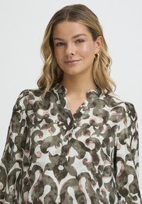 Gombos blúz zöld és rózsaszín absztrakt mintával fehér alapon, álló gallérral és texturált anyagból.