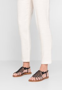 Sandales noires en cuir avec un design tissé et une sangle ajustable à la cheville, montées sur une semelle en bois brun clair, portées avec un pantalon crème.