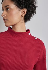 Czerwony sweter z prążkowanego materiału i wysokim kołnierzem, z akcentami w postaci srebrnych guzików na ramieniu. Tkanina wydaje się miękka i fakturowana.