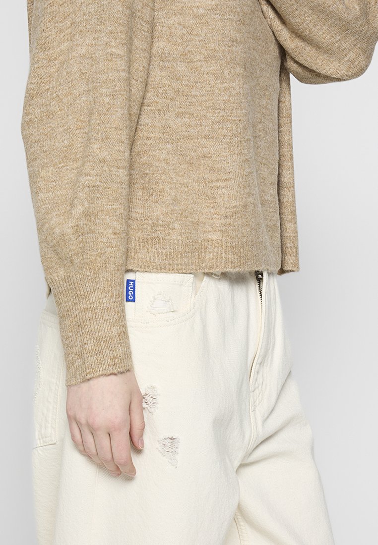 Suéter de punto beige con mangas largas, con un corte holgado y puños acanalados, combinado con pantalones color crema que tienen detalles desgastados y una etiqueta de logo.