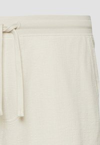 Gros plan sur la taille d'un pantalon en tricot couleur crème, avec un cordon de serrage noué en un nœud lâche sur un fond clair.