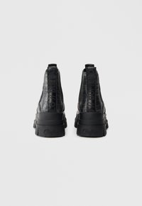 Μαύρα μποτάκια αστραγάλου με υφή κροκοδείλου, με χοντρή σόλα και τακούνι με ανάγλυφο λογότυπο. Σχεδιασμένα χωρίς κορδόνια για εύκολη εφαρμογή.