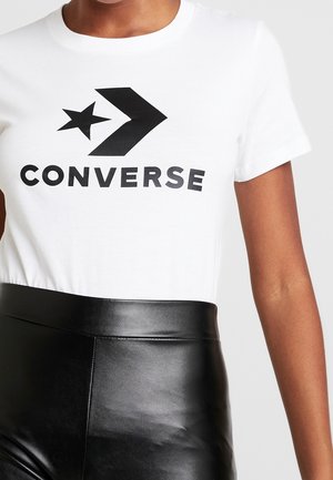 Gros plan d'une personne portant un t-shirt blanc avec le logo noir Converse et un pantalon en cuir noir taille haute.
