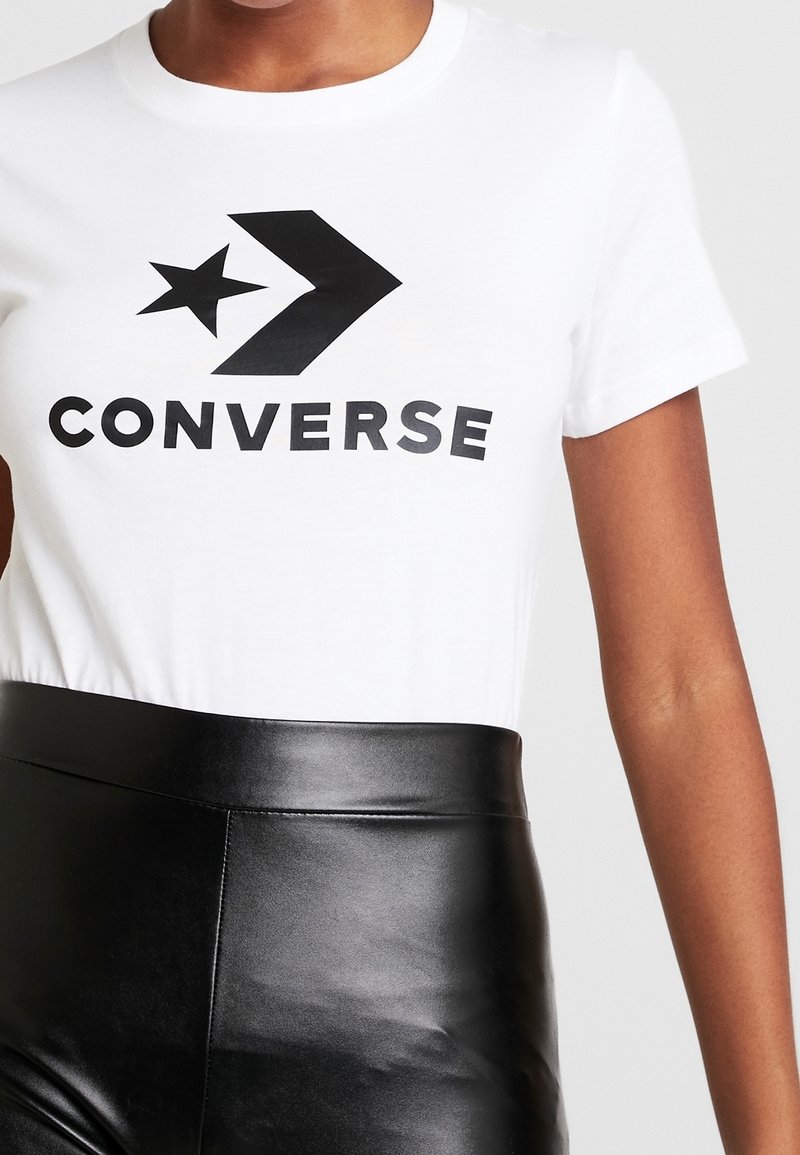 Gros plan d'une personne portant un t-shirt blanc avec le logo noir Converse et un pantalon en cuir noir taille haute.