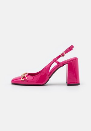 Błyszczący różowy sandał na wysokim obcasie typu slingback z detalem złotej klamry w kształcie serca i grubym obcasem, pokazany z boku na białym tle.