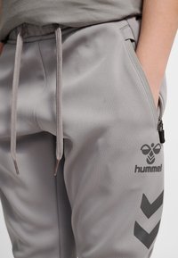 Osoba nosząca jasnoszare spodnie dresowe Hummel z sznurkiem w pasie i zamykaną na zamek błyskawiczny boczną kieszenią, ręka umieszczona w kieszeni.