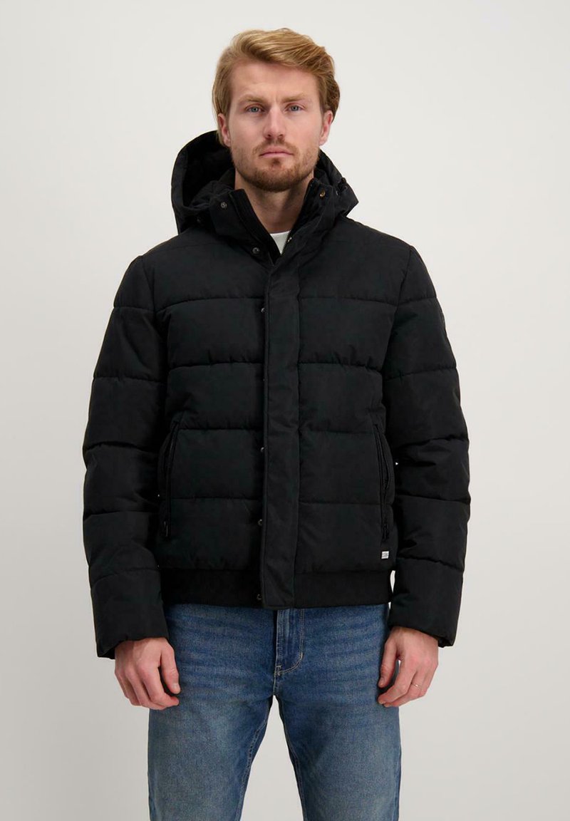 Zwarte pufferjack met een rits- en drukknoopsluiting, met een capuchon, zijzakken en een gequilt textuur. Ggedragen met een blauwe broek.