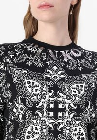Svart jersey-topp med hvitt paisley-mønster og blomstrende motiver. Skinnende "PINKO"-tekstdetalj nær halsen. Korte ermer.