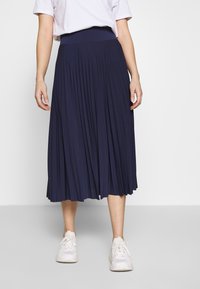 Mörkblå veckad kjol med en bred elastisk midja, tillverkad av mjukt material. Bärs med vita sneakers som har en slät yta och ett flödande design.