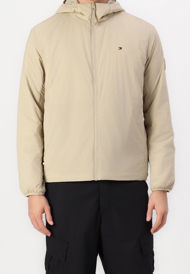 Beżowa kurtka zapinana na zamek z kapturem, elastycznymi ściągaczami na rękawach i małym logo na piersi, noszona z czarnymi spodniami typu cargo.