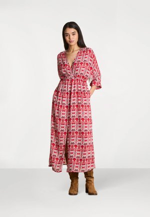 Femme debout, les mains dans les poches, portant une longue robe rouge à motifs et des bottines à talons en daim marron sur un fond blanc uni.