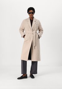 Cappotto in lana beige con cintura, colletto a rever, tasche laterali, abbinato a pantaloni grigi e scarpe slip-on nere. Modello che indossa occhiali da sole.