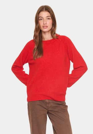 Roter Strickpullover mit Rundhalsausschnitt, langen Ärmeln und gerippten Bündchen sowie Saum, kombiniert mit braunen Hosen. Weiche Textur und entspannte Passform.