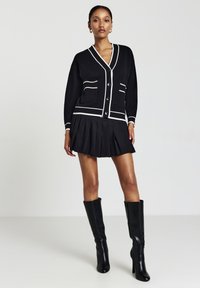 Femme portant un cardigan noir avec des bordures blanches, une mini-jupe plissée noire et des bottes noires à hauteur de genoux, se tenant contre un fond uni.