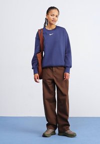 Sudadera navy con escote redondo y logo, pantalones sueltos marrones y zapatillas verdes. El modelo sostiene un bolso marrón cruzado sobre el cuerpo.
