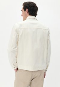 Jachetă din denim albă cu un guler clasic, manșete cu nasturi și o croială dreaptă. Textură netedă și design minimalist, cu cusături pe spate.