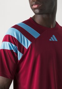 Rotes Sporttrikot mit hellblauen diagonalen Streifen auf den Schultern, gefertigt aus einem glatten Stoff, mit Rundhalsausschnitt und Logodetail.