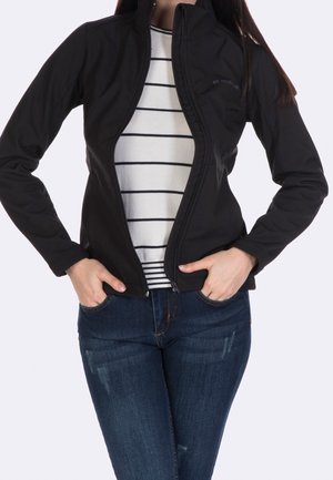 Woman wearing an unzipped black jacket over a white long-sleeve shirt with black horizontal stripes and blue jeans, hands in jacket pockets.