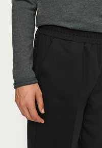 Detailní záběr na ruku osoby, která odpočívá vedle černých kalhot s elastickým pasem a šedého pleteného topu s dlouhým rukávem a ohrnutými manžetami.