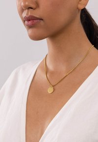 Goldkette mit rundem Anhänger, versehen mit einer zarten Kette, getragen über einem tiefen V-Ausschnitt in Weiß. Schlichte und glatte Ausführung.