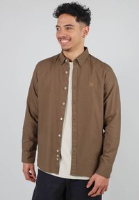 Chemise boutonnée marron avec un col, en coton, ornée d'une broderie sur la poitrine. Portée par-dessus un t-shirt de couleur claire.