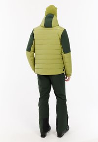 Zelena izolirana jakna s kapuljačom, sa šivenim dizajnom. Uparena s tamnozelenim hlačama i crnim čizmama, minimalno vidljivo brendiranje.