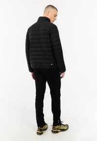 Vyras, vilkintis juodą siūtu striukę, juodas kelnes ir geltonus žygių batus, stovi ir žiūri per petį priešais baltą foną.