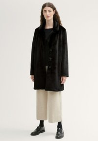 Cappotto in pelliccia sintetica nera con ampio colletto, bottoni frontali e maniche lunghe. Indossato sopra pantaloni cropped beige chiaro e scarpe nere.