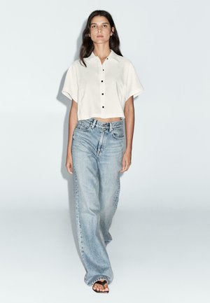 Chemise blanche cropped avec boutons noirs, associée à un jean bleu clair ample. Sandales plates complètent la tenue. Tissu lisse et coupe décontractée.