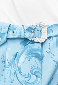 Ανοιχτό γαλάζια ζώνη από ύφασμα με μοτίβο, ασημένιες οπές και περίτεχνη λευκή αγκράφα με ανάγλυφο το κείμενο "Versace Jeans Couture".