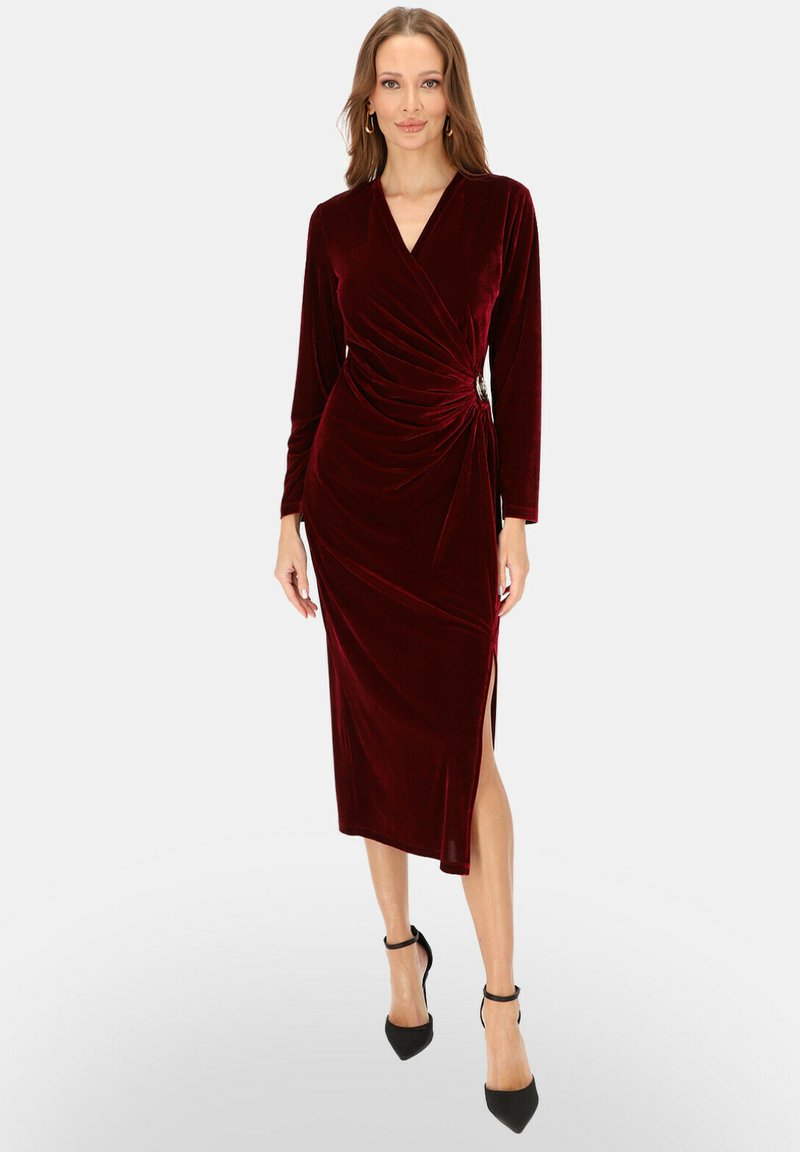 Burgundowa welurowa sukienka kopertowa z długimi rękawami, bocznym rozcięciem i marszczeniem w talii, ozdobiona dekoracyjną przypinką. Czarne szpilki.