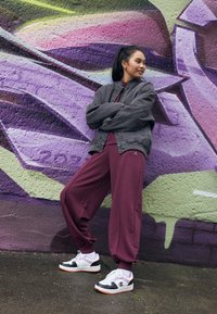 Burgundowe dresy i szara, luźna kurtka, w połączeniu z czarno-białymi sneakersami. W tle znajduje się żywe fioletowe graffiti.