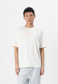 T-shirt en coton couleur crème avec une coupe décontractée, des manches courtes et un logo "Hollister" rose sur la poitrine. Design simple et texture lisse.