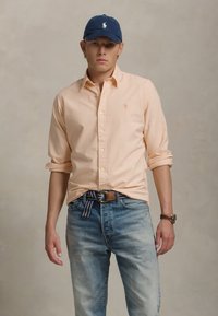 Ανοιχτό πορτοκαλί πουκάμισο με κουμπιά, ανοιχτό μπλε τζιν, μπλε καπέλο με λογότυπο, καφέ ζώνη με ριγέ λεπτομέρεια, καφέ ρολόι. Απλό φόντο.