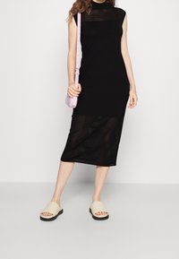 Robe noire sans manches à rayures verticales, longueur au genou. Associée à des chaussures beige à enfiler et un sac bandoulière rose clair. Design simple.