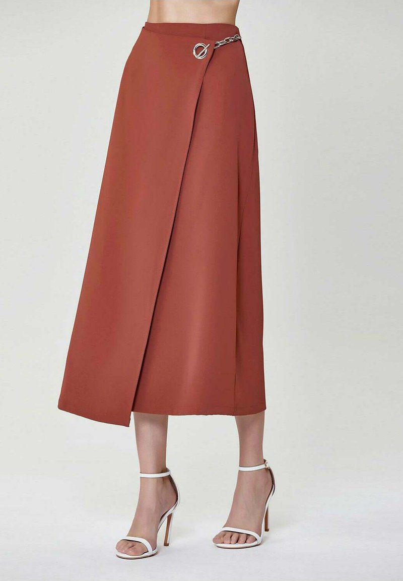 Jupe midi couleur rouille avec un design enveloppant, présentant un accent circulaire argenté et un détail de chaîne à la taille. Texture lisse.