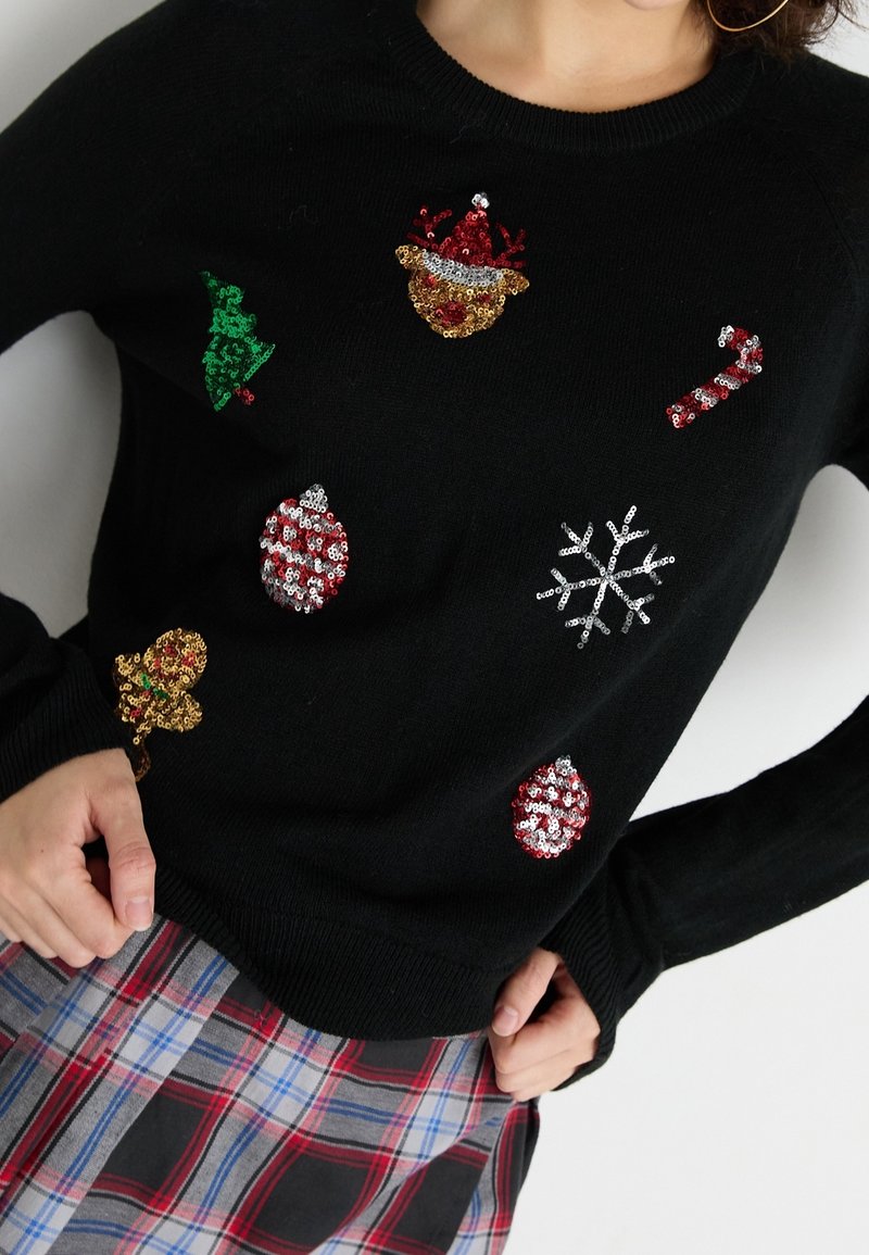 Pull noir avec motifs de Noël pailletés comprenant arbre, bonnet du Père Noël, canne en sucre d'orge, flocon de neige et bonhomme en pain d'épices, assorti à une jupe à carreaux.