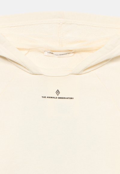 Εγγύς άποψη του λαιμού μιας κρεμ-χρωματισμένης κουκούλας με το κείμενο "THE ANIMALS OBSERVATORY" και το λογότυπο τυπωμένα κάτω από το γιακά, μέσα και έξω από το ένδυμα.