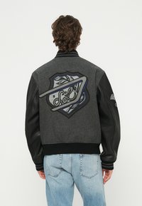 Osoba s kučeravými hnedými vlasmi, oblečená v sivo-čiernej vysokoškolskej bunda s veľkou vyšívanou nášivkou štítu na chrbte, v kombinácii so svetlomodrými džínsami.